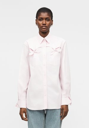 Nainen, jolla on päällään vaaleanpunainen nappipaita, jossa on koristeelliset rusetit rintataskujen yläpuolella, yhdistettynä vaaleansinisiin farkkuihin.