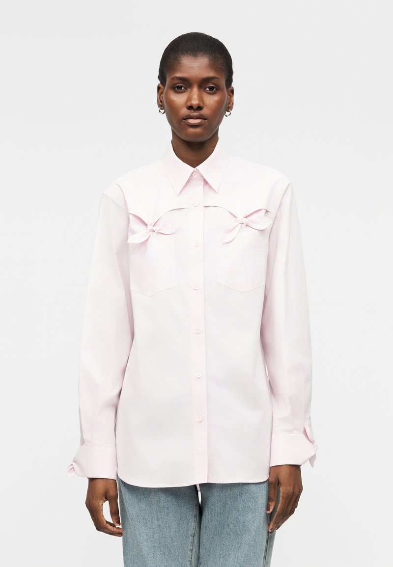 Femme portant une chemise rose pâle boutonnée avec des nœuds décoratifs au-dessus des poches poitrine, associée à un jean bleu clair.