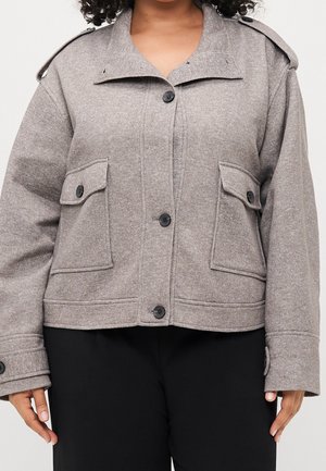 Veste grise boutonnée avec poches à rabat et épaulettes portée sur un pantalon noir par une personne aux cheveux bouclés.