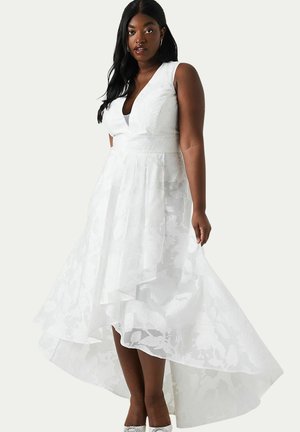 Femme portant une robe blanche sans manches à texture florale, coupe asymétrique, avec un décolleté en V profond et des talons à bride à la cheville.