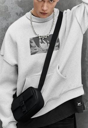 Giovane persona che indossa una felpa con cappuccio grigio chiaro con grafica anime, collana a catena d'argento, borsa a tracolla nera e maglietta nera su sfondo grigio texturizzato.