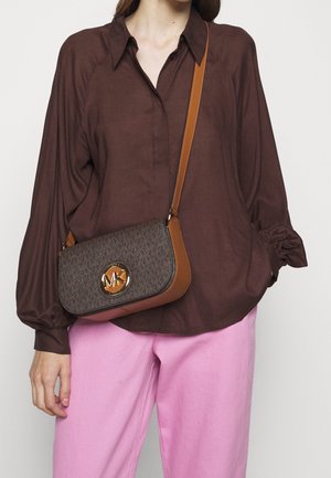 Brun crossover taske med monogrammønster, guldhardware og en tan læderdetalje; båret med en løs brun bluse og lyserøde bukser.