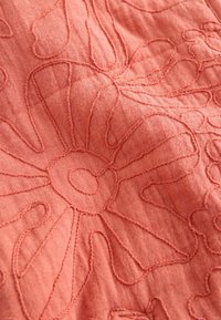 Közelkép korallszínű anyagról, melyen kiemelkedő virágos hímzés található, nagy virágmintákat alkotva.