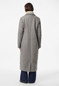 Žena v dlouhém šedém vlněném kabátu, modrých širokých džínách a černých botách, stojící zády k fotoaparátu před jednobarevným bílým pozadím.