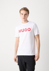 Bílá bavlněná tričko s výrazným červeným logem "HUGO". Krátké rukávy a kulatý výstřih; hladká textura a ležérní střih.
