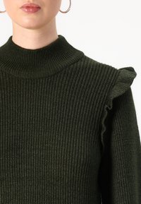 Pull en laine côtelée vert foncé avec un col haut et des accents froncés sur les épaules. La texture en tricot ajoute de la chaleur et de la dimension au design.