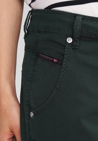 Pantalons en denim vert foncé avec une fermeture éclair, des coutures décoratives et une étiquette de marque visible sur la poche avant. Accents en matériel argenté.