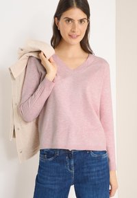 Suéter de color rosa claro con cuello en V, hecho de una tela suave, que presenta un ajuste relajado y mangas largas. Combinado con jeans de mezclilla azul oscuro.