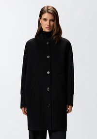 Cappotto di lana nero con colletto alto, chiusura frontale con bottoni, due tasche laterali e polsini a costine. Silhouette oversize e strutturata.