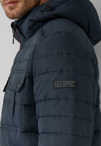 Tumesinise puffer-jakk kapuutsi ja kaheosalise kudumisega, esitaskud ja bränditud nahapatch varrukal.