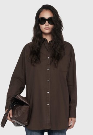 Woman with long dark hair wearing black sunglasses, a loose dark brown button-up shirt, holding a large dark brown leather bag.