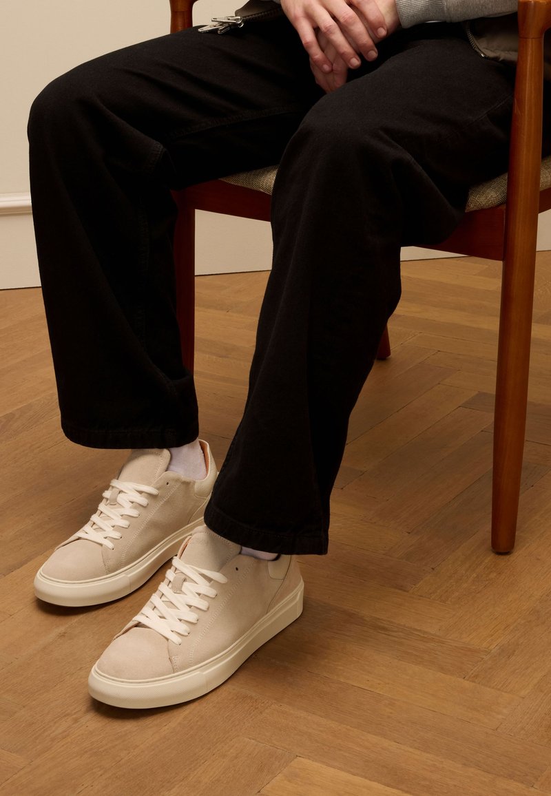 Beige Wildlederschuhe mit weißen Schnürsenkeln, runder Zehenpartie und einer glatten Gummisohle, getragen mit dunklen Hosen, sitzend auf einem Holzstuhl.