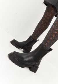 Botas de cuero negras hasta el tobillo con un frente texturizado, cremallera lateral, suela gruesa y una pestaña para tirar en la parte posterior. Llevadas con pantis negros con estampado.