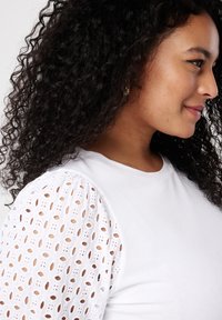 Weißes T-Shirt mit kurzen Puffärmeln und aufwendiger Lochstickerei, das ein strukturiertes Design auf einem glatten Stoff zeigt.