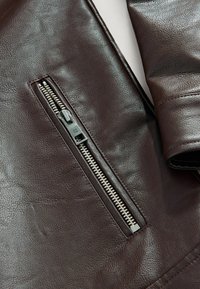 Manche de veste en cuir brun foncé avec une poche zippée verticale en argent et une texture de cuir visible sur un fond clair.