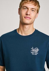 Námornícky modré tričko s krátkym rukávom vyrobené z bavlny, s grafickým logom ryby a hranolkov na ľavej strane hrude.