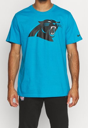 Homme portant un t-shirt bleu vif avec un logo noir de panthère rugissante et un pantalon noir avec un petit patch logo NFL visible.