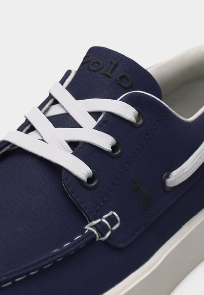 Μπλε σκούρο καμβά παπούτσι για σκάφος με λευκά κορδόνια, λευκή σόλα και τη λέξη «Polo» στο γλώσσα και την πλευρά.