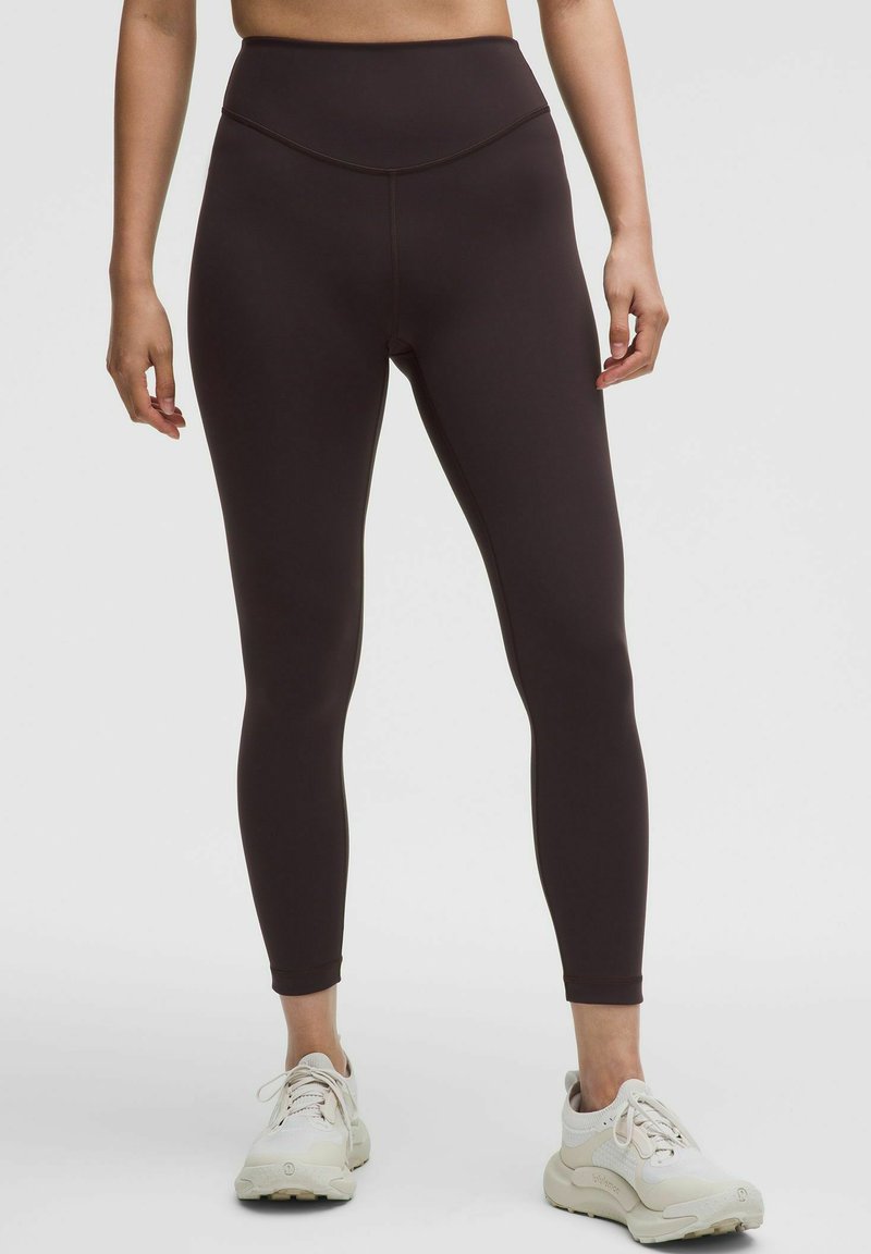 Person wearing high-waisted dark brown leggings and white athletic shoes, standing against a plain light background.