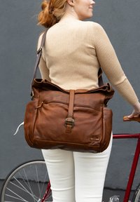 Brūna ādas sūtītāja soma ar pārklāju, regulējamu siksnu un priekšējo kabatu. Iespējas ietver teksturētu apdari un misiņa detaļas.