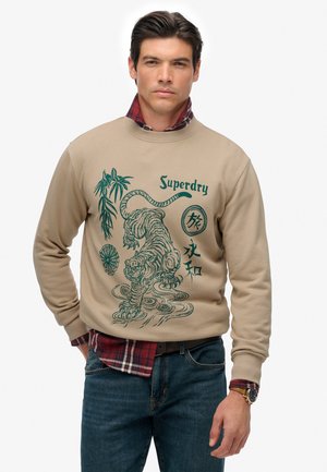Homme portant un sweat beige avec un design de tigre vert et de bambou, une chemise à carreaux rouges en dessous, un jean bleu et une montre au poignet gauche.