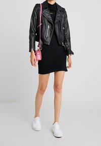 Schwarze Lederbikerjacke, eng anliegendes schwarzes Kleid, pinke Handtasche und weiße Sneakers. Auffälliger Ausschnitt im Kleid und strukturierte Jackenoberfläche.
