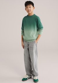 Pull ombré vert avec col côtelé, associé à un jean baggy gris clair et des baskets avec des accents verts. Enfant debout, mains dans les poches.