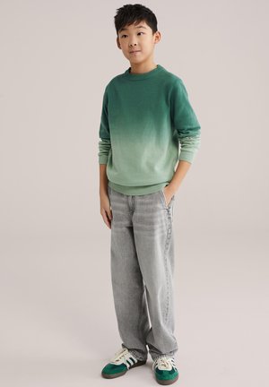 Grüner Ombre-Pullover mit geripptem Kragen, kombiniert mit hellgrauen, weit geschnittenen Jeans und Sneakers mit grünen Akzenten. Kind steht, Hände in den Taschen.