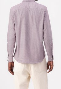 Camisa de manga larga a cuadros rojos y blancos con dobladillo curvado, puños con botón y detalle en la yugo trasero. Combinada con pantalones de color claro.