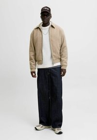 Chaqueta bomber color bronce, suéter de punto blanco, pantalones de denim oscuros y sueltos, y zapatillas beige. La chaqueta tiene cuello y cremallera al frente; gorra casual.