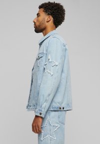 Light blue denim jacket featuring star-shaped distressing on sleeves, buttoned front, and slight fabric fading with white paint splatters.