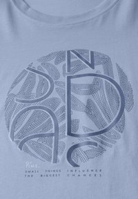 Camiseta de algodón azul claro con un diseño circular texturizado que presenta texto estilizado y patrones de hojas en un azul más oscuro.