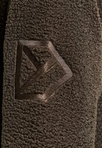 Brauner Fleece-Stoff mit strukturierter Oberfläche und einem dunkelbraunen geometrischen Logo, das an der Seite eingeprägt ist.