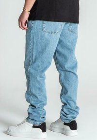 Jeans de denim azul deslavado con un corte relajado, que presentan dos bolsillos traseros, combinados con zapatillas blancas y negras. Textura suave y diseño casual.