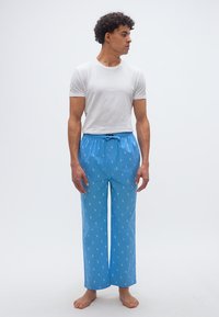 Άνδρας που στέκεται ξυπόλυτος, φοράει λευκό κοντομάνικο μπλουζάκι και ανοιχτό μπλε παντελόνι πυτζάμας με μικρά λευκά σχέδια, κοιτάζοντας προς το πλάι.