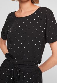 Haut noir à manches courtes avec un col dégagé, présentant un motif à pois blancs. Le tissu semble lisse avec une texture subtile, assorti à un pantalon noir.