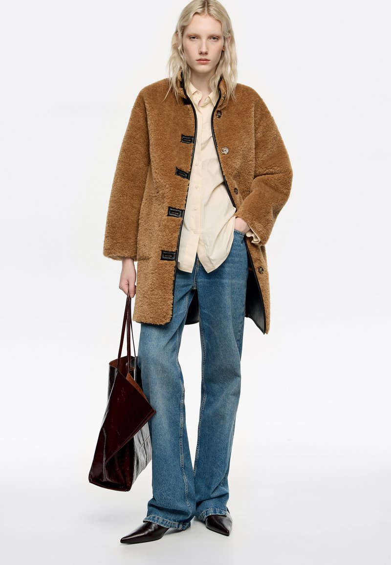 Mujer joven llevando un abrigo de borrego marrón, una camisa beige, jeans azules y zapatos de punta negros, sosteniendo un gran bolso de cuero marrón oscuro.