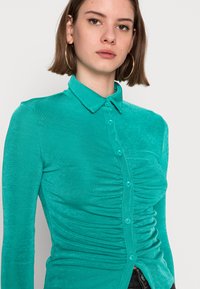 Camisa ajustada turquesa com mangas longas, frente abotoada e textura franzida nas laterais. Apresenta um colarinho clássico e botões combinando.