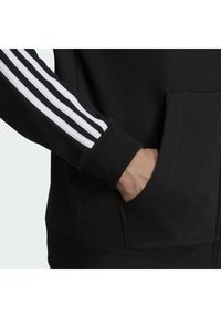 La mano di una persona nella tasca a canguro di una felpa nera con tre strisce bianche sul polsino delle maniche.