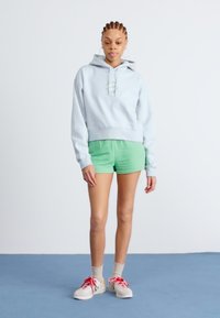 Světle modrá zkrácená mikina s kapucí a šňůrkami, spárována se zelenými kraťasy. Model má na sobě bílé boty s červenými akcenty a kotníkové ponožky na modré podlaze.