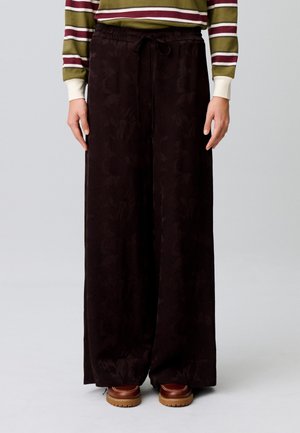 Persona che indossa pantaloni neri larghi e morbidi con coulisse in vita, top a righe a maniche lunghe e scarpe marroni, in piedi su sfondo chiaro.
