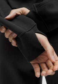 Sudadera negra con puños acanalados, mostrando un primer plano de manos sujetando la tela. La textura parece suave y elástica.