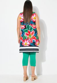 Kolorowa bezrękawnikowa tunika z dużym kwiatowym wzorem w odcieniach różu i zieleni, połączona z zielonymi legginsami 3/4 i beżowymi butami na platformie.