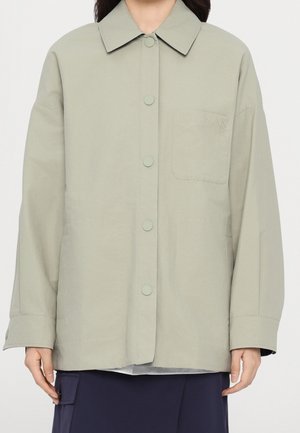 Personne portant une veste-chemise gris clair boutonnée avec une poche poitrine et un pantalon foncé, montrée du cou aux hanches.