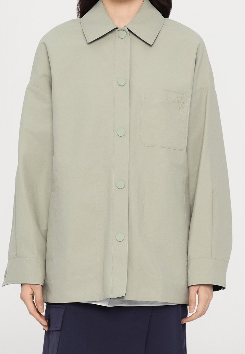 Personne portant une veste-chemise gris clair boutonnée avec une poche poitrine et un pantalon foncé, montrée du cou aux hanches.