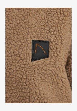 Beige Fleece-Stoff mit einer strukturierten Oberfläche, versehen mit einem schwarzen Patch, der ein orangefarbenes geometrisches Design zeigt. Vollzip-Design abgebildet.