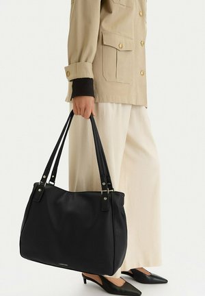 Ręka trzymająca dużą czarną skórzaną torbę na ramię z podwójnymi uchwytami, ubrana w beżową kurtkę, białe szerokie spodnie i czarne buty na obcasie.