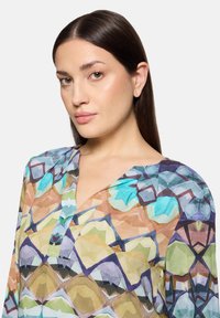 Kolorowa, przezroczysta bluzka z geometrycznymi wzorami, z delikatnym dekoltem w serek i miękką fakturą tkaniny. Dominujące kolory to niebieski, zielony i brązowy.