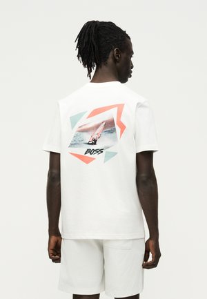 Homme portant un t-shirt blanc avec un motif coloré de voilier et le texte "BOSS" dans le dos, associé à un short blanc, debout de dos.