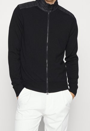 Mand iført en sort jakke med lynlås og polstrede skuldre samt hvide bukser, hænderne i lommerne, foran en ensartet lys baggrund.